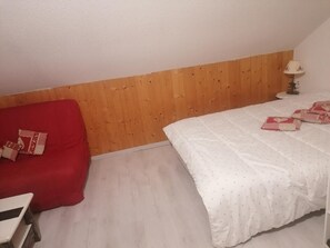 Ein Schlafzimmer mit einem hölzernen Kopfteil, einem roten Schlafsofa, einem weißen Bett mit gemusterten Betttextilien und einer Nachttischlampe.