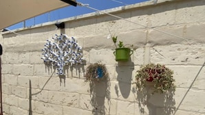 Una terraza con un muro blanco, tres macetas colgantes con plantas y un cuadro eléctrico a la vista.