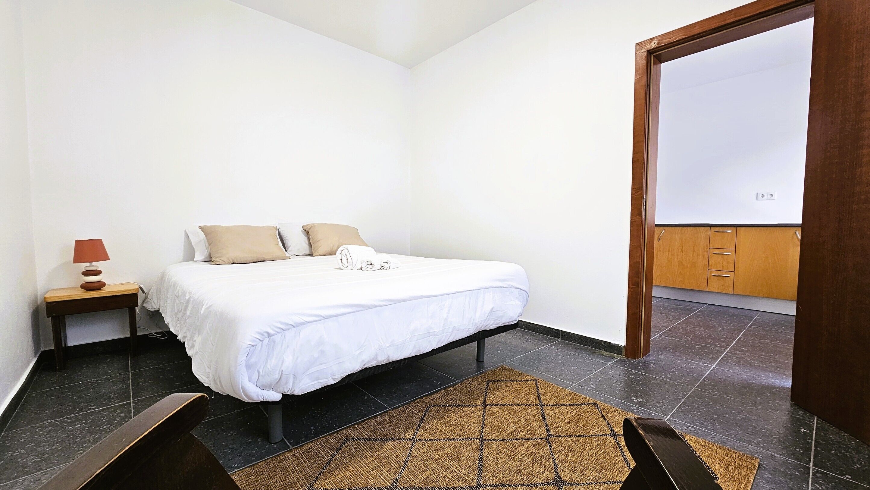A bedroom with a bed, bedside table, lamp, and a view of the outside.