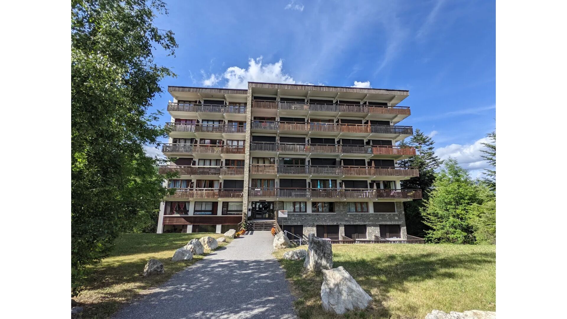 Een hotel met meerdere verdiepingen, balkons, een centrale ingang en een omliggend natuurlandschap.