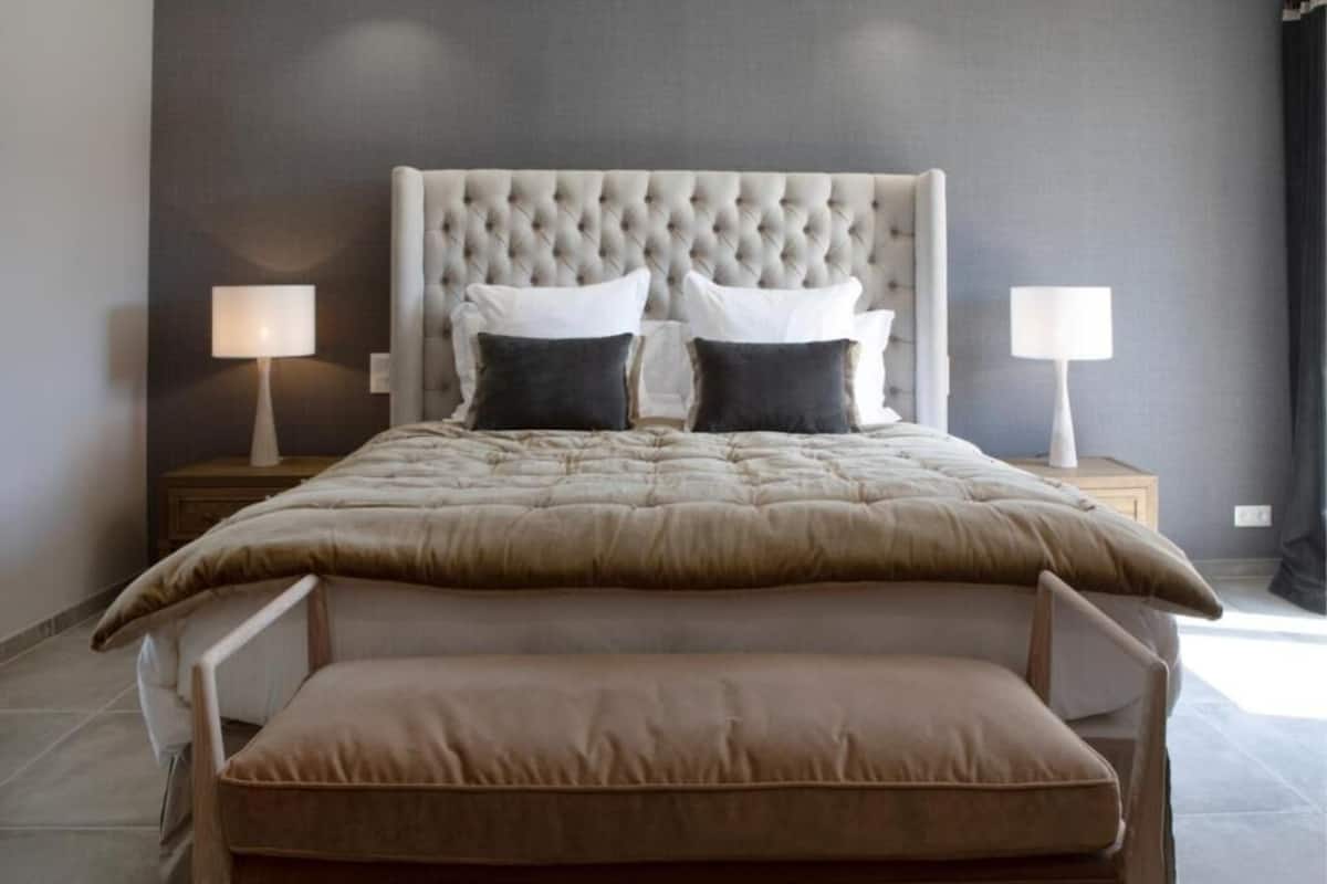 Camera da letto con testiera trapuntata, un letto grande con copripiumino color beige, due lampade da comodino e un comodino in legno.