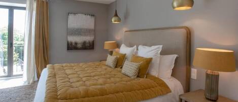 A large bed with a yellow duvet, two bedside lamps, a painting on the wall, and a view through a window.