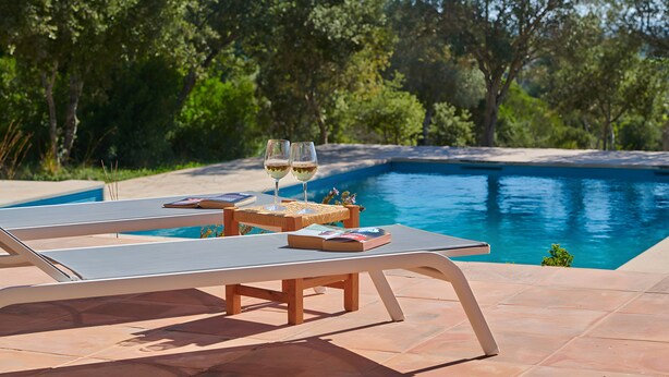 A poolside area with lounge chairs and a table, two glasses of wine, and a book. - Country House 'Fetget' with Mountain View, Private Pool and Wi-Fi (Illes Balears)
