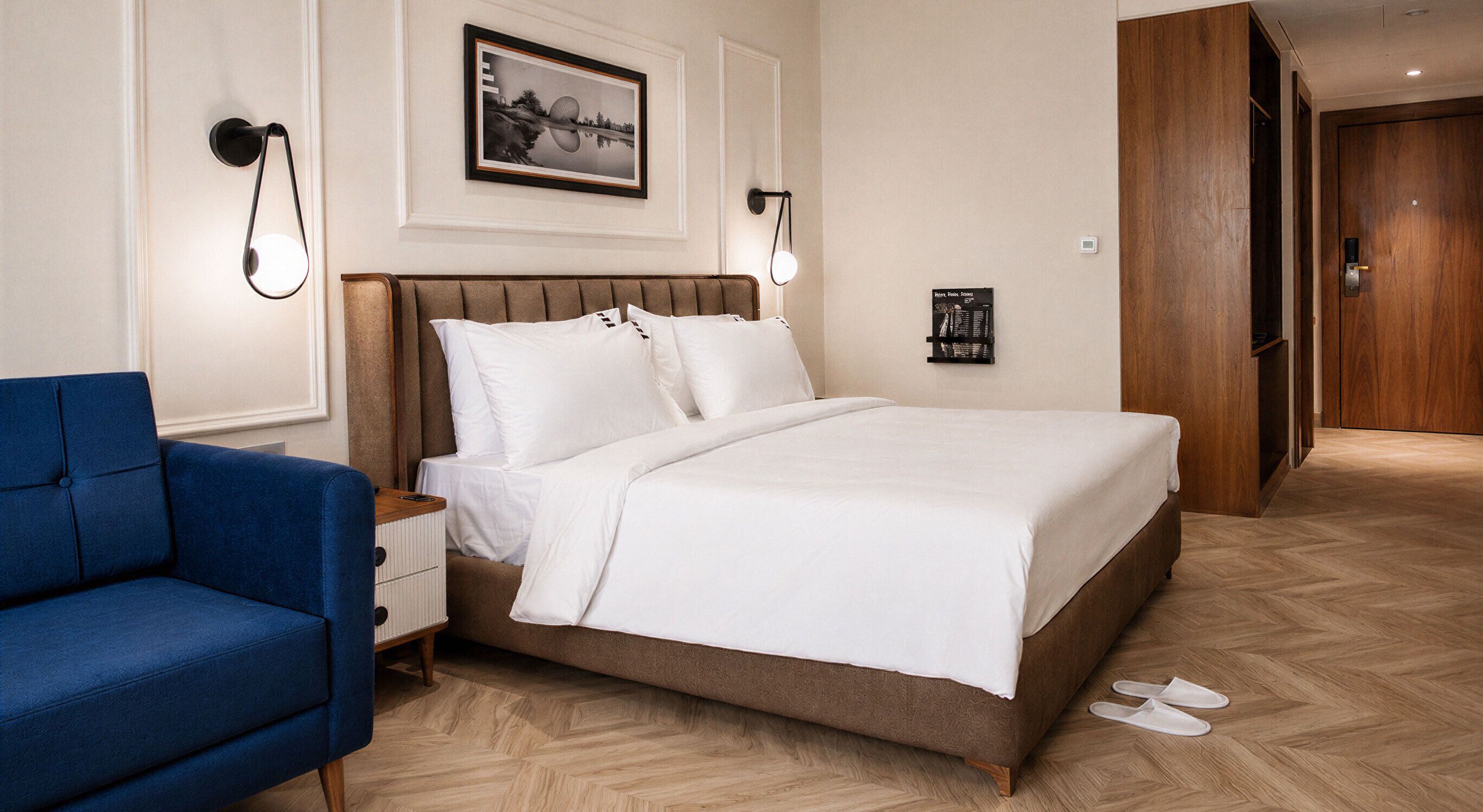 A hotel room with a bed, a blue armchair, a nightstand, a wall-mounted lamp, a framed picture, and a wooden door.