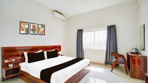 A hotel room with a large bed, a bedside table with a lamp, a desk with a chair, and a window with curtains. - Deluxe Hebbal Townhouse (Bengaluru)