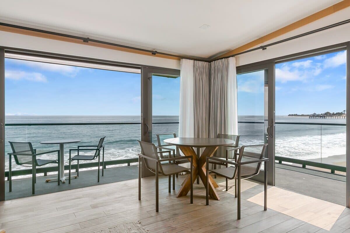 A room with a round table and chairs, large windows with a view of the sea, and a balcony with a railing.