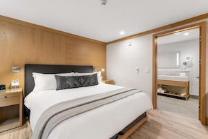 Una habitación de hotel moderna con una cama con cabecera de madera, una mesita de noche con lámpara, y un baño con espejo y lavabo.