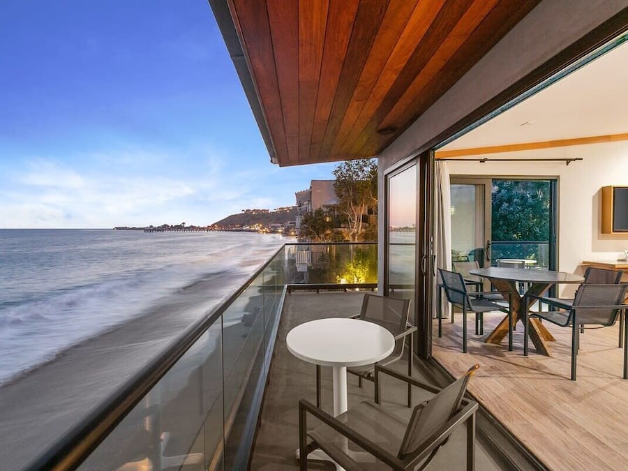 Un balcon moderne avec une balustrade en verre donnant sur l’océan, une table ronde avec des chaises et un téléviseur à écran plat fixé au mur.