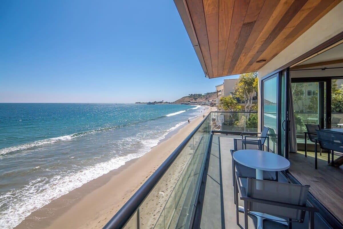 A balcony with a view of the beach and ocean, a table, and chairs.