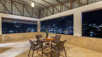 A rooftop terrace with a round table, chairs, and a view of a cityscape at night.