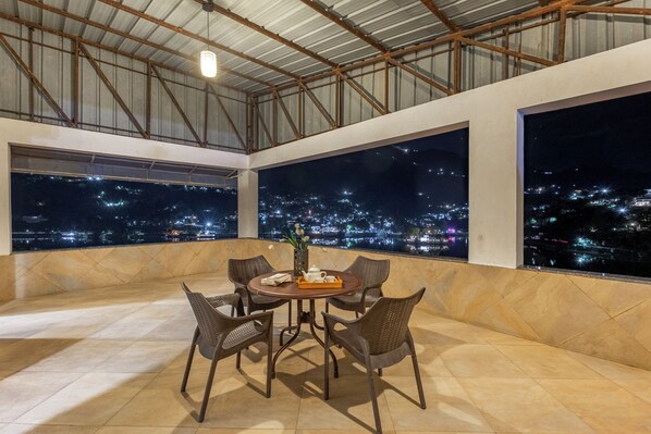 Una terraza en la azotea con una mesa redonda, sillas y vistas a un paisaje urbano nocturno.