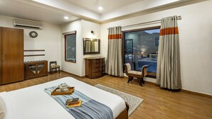 A hotel room with a bed, a desk, a chair, a wardrobe, an air conditioner, a clock, a window with curtains, and a view of the cityscape. - ELIVAAS Lakeview Manor 5 BHK  (Bhimtal)