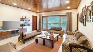 A spacious living room with a wooden ceiling, a large window with a view, a flat-screen TV, and a sofa set. - ELIVAAS Lakeview Manor 5 BHK  (Bhimtal)
