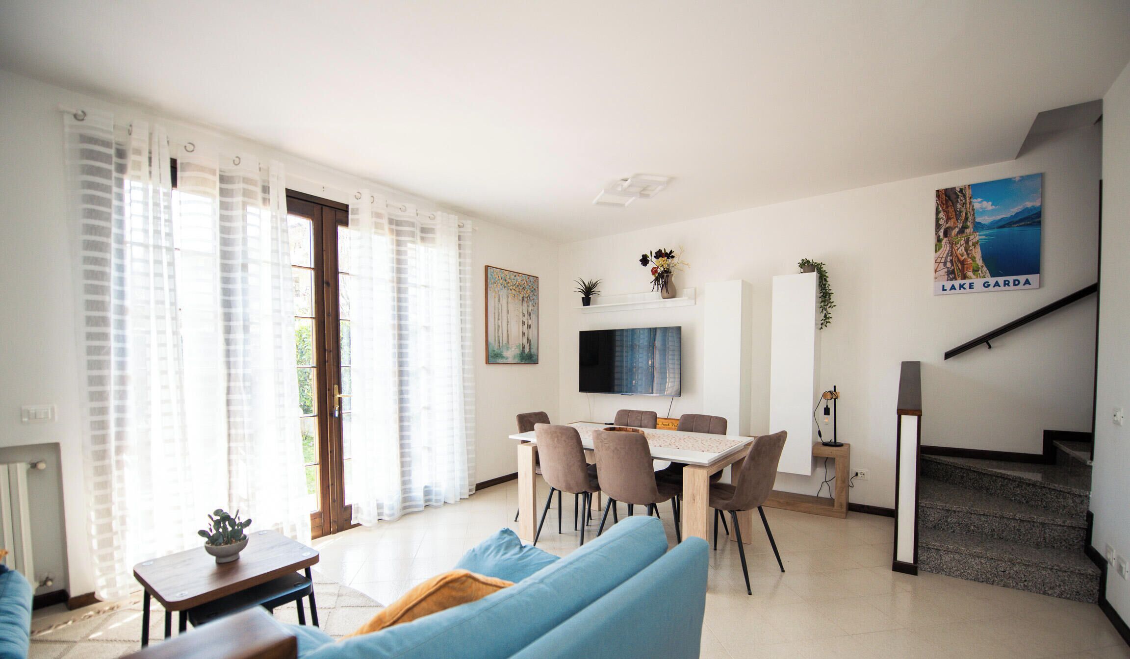 Uma sala de estar moderna com área de jantar, escada e uma grande janela com cortinas leves.