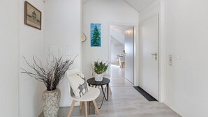 Un pasillo con pared blanca, un cuadro enmarco, una planta en maceta, una silla con un cojín de ciervo y una mesita con otra planta.