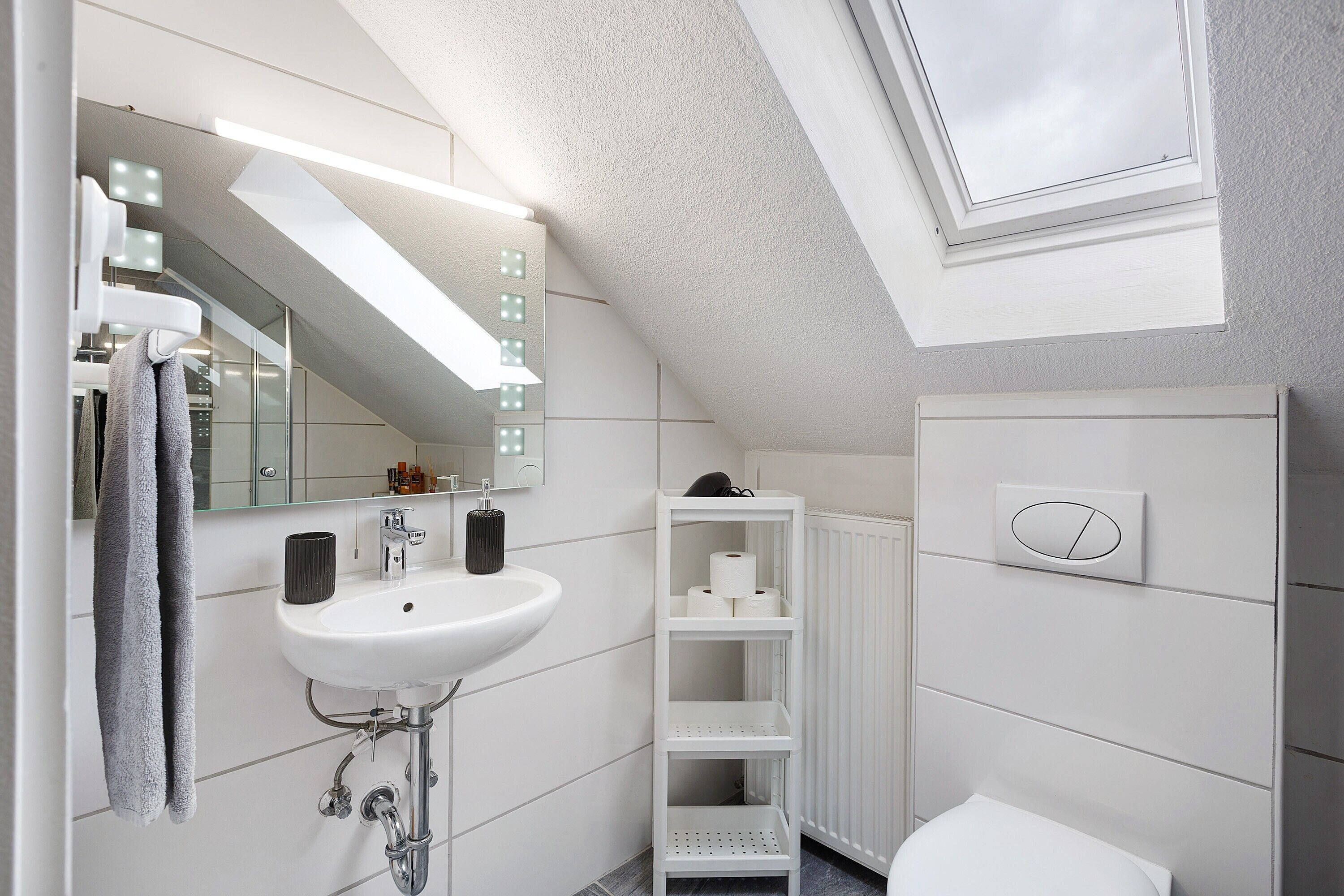 Un baño moderno con tragaluz, un lavamanos blanco, un espejo con iluminación, un perchero para toallas y un inodoro.