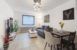 A modern living room with a sofa, coffee table, dining table, chairs, a radiator, a TV, a plant, and artwork on the walls. - Schwarzwaldglück – Group Getaway with Balcony & Garden (2 Apartments) (Sankt Blasien)