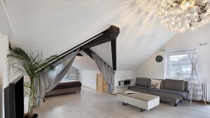 Un dormitorio tipo loft moderno con techo inclinado, cama empotrada, sofá, mesa de centro, televisor y una planta grande en maceta.