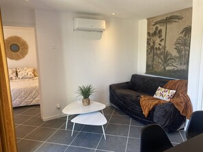 A living room with a dark sofa, a small white table, a potted plant, and a painting on the wall. - Ground floor apartment, AC and parking - 4 guests (Le Barcarès)