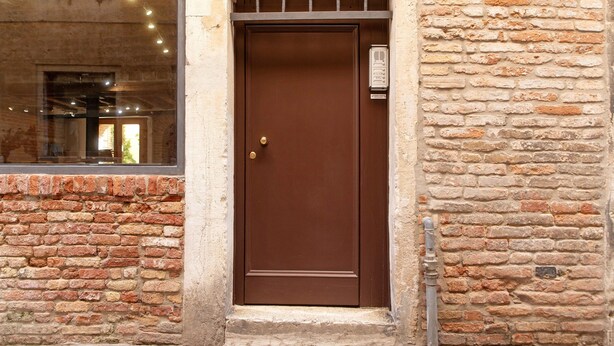 Una puerta marrón con manija de latón, una ventana que da a un espacio interior y una pared de ladrillo.
