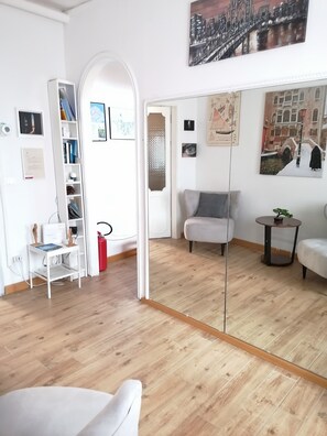 Ein Zimmer mit Holzboden, einem weißen Sofa, einem kleinen Tisch mit Pflanze, einem Bücherregal und einer Tür zu einem anderen Raum.