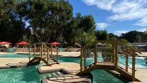 A swimming pool with wooden bridges and steps, surrounded by trees and umbrellas.