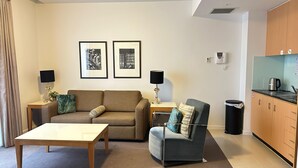 A living room with a sofa, armchair, coffee table, and a small side table with a lamp. There are framed pictures on the wall, a kitchen area with a kettle, and a trash bin. - The Heritage Bank Apartment (Melbourne)