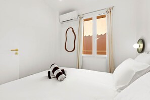 A hotel room with a bed, white linens, a window with curtains, an air conditioning unit, and a wall-mounted light fixture. - Exceptional house with sea view and pool (Roquebrune-sur-Argens)