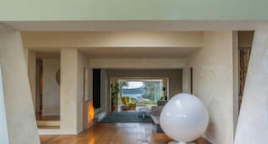 Een modern interieur met een houten vloer, een groot wit bolvormig object, en een uitzicht door een deuropening op een buitenruimte met planten en een stoel.