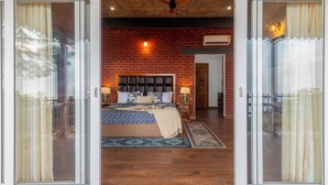 A bedroom with a brick wall, a bed with a headboard, two bedside tables with lamps, and a sliding glass door leading to a balcony. - ELIVAAS Fleur De Lis 4 BHK Villa (Kasauli)