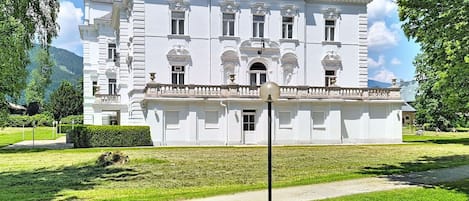 Un edificio grande y blanco de estilo clásico con numerosas ventanas y una entrada central, rodeado de céspedes cuidados y árboles.