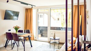 Un saló modern amb un sofà, una taula de menjador amb caires, un televisor fixat a la paret i una vista per la porta de vidre.