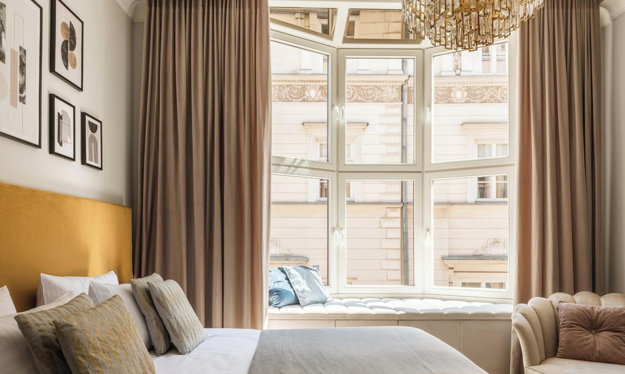 Ein Hotelzimmer mit großem Fenster, einem Bett mit Kissen, einer Couch und gerahmten Kunstwerken an der Wand.