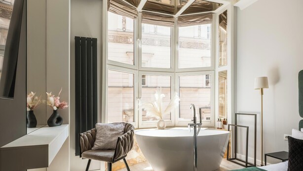 Ein modernes Badezimmer mit einer freistehenden Badewanne, einem Sessel und einem großen Fenster mit Blick auf Gebäude.