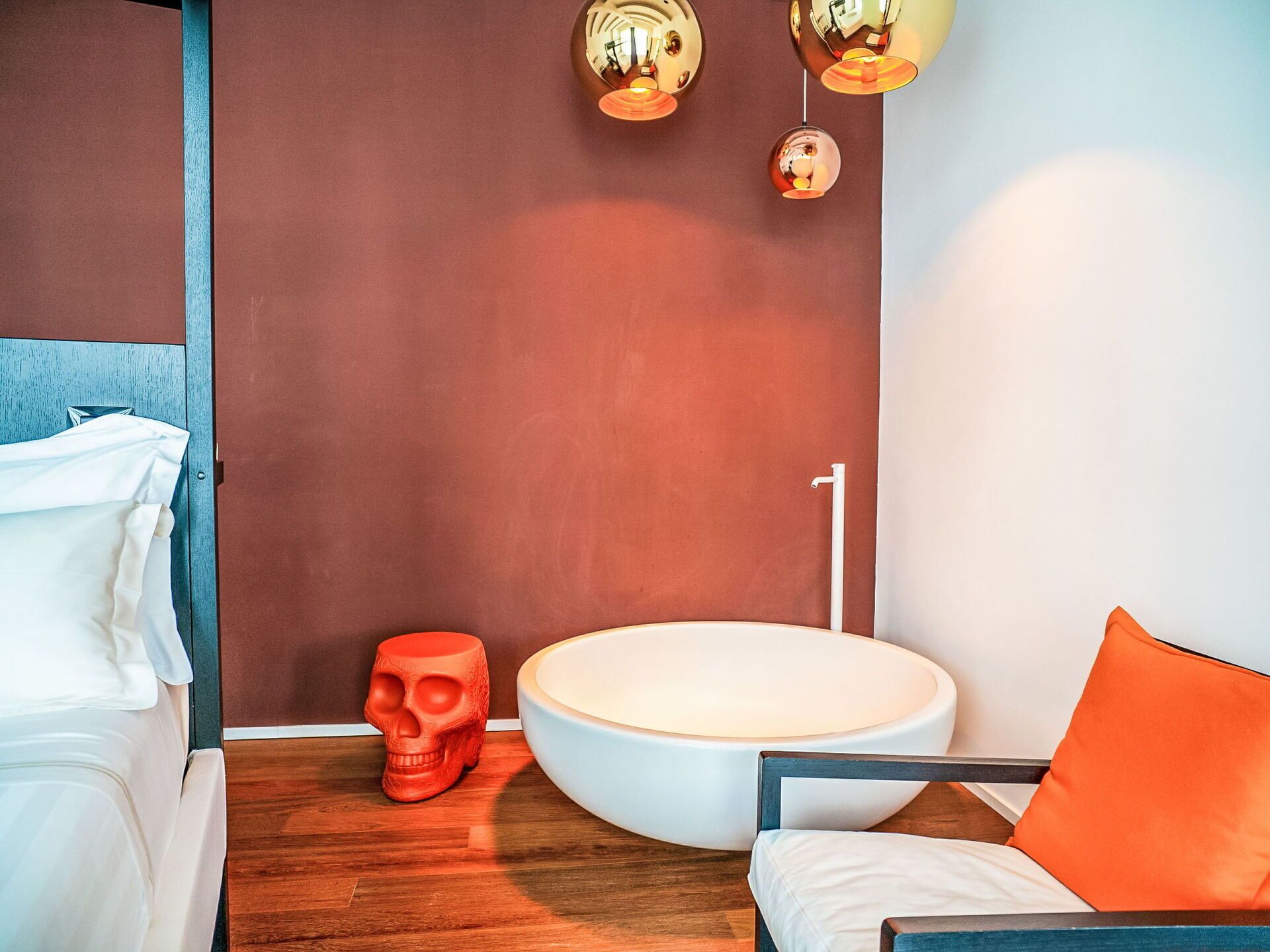 A modern bathroom with a wooden floor, a white bathtub, a red skull decoration, and a white chair with an orange pillow.
