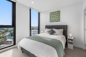 A modern bedroom with a large bed, a nightstand, a bedside table, a window with a view, and a green floral artwork on the wall. - Level 9 Views · 5 min to Austin Hospital | Parking (Ivanhoe)