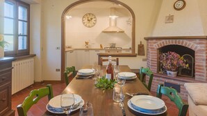 Comedor con una mesa de madera dispuesta para cuatro, una chimenea de ladrillo y una cocina al fondo.