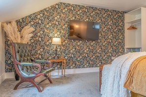 A bedroom with a patterned wallpaper, a flat-screen TV, a tufted leather chair, a bedside table, and a bed with a white cover. - Gorgeous 3 Bed House Lancashire (Morecambe)