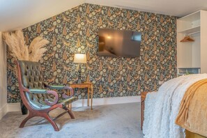 A room with a patterned wallpaper, a television mounted on the wall, a chair, a small table, and a bed with a white blanket. - Gorgeous 3 Bed House Lancashire (Morecambe)