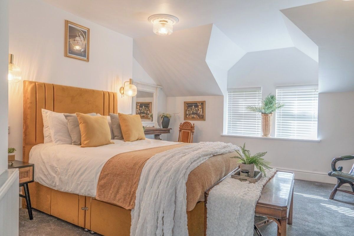 A bedroom with a large bed, a bench, a desk, and a window with blinds.