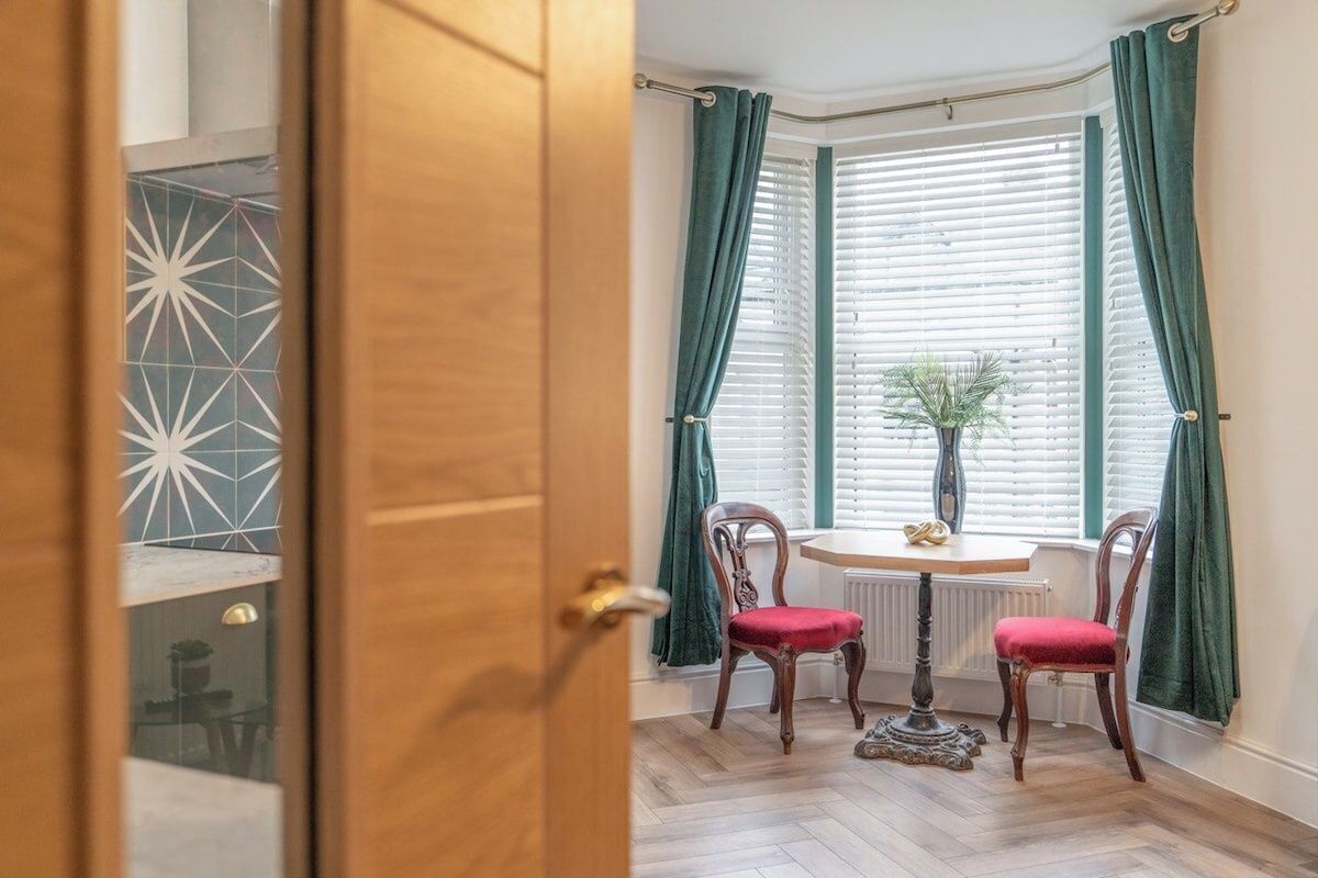 A room with a wooden door, a small table with chairs, a radiator, and a window with curtains.