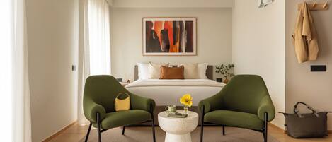 A modern bedroom with a bed, two green armchairs, a round coffee table, and a painting on the wall.