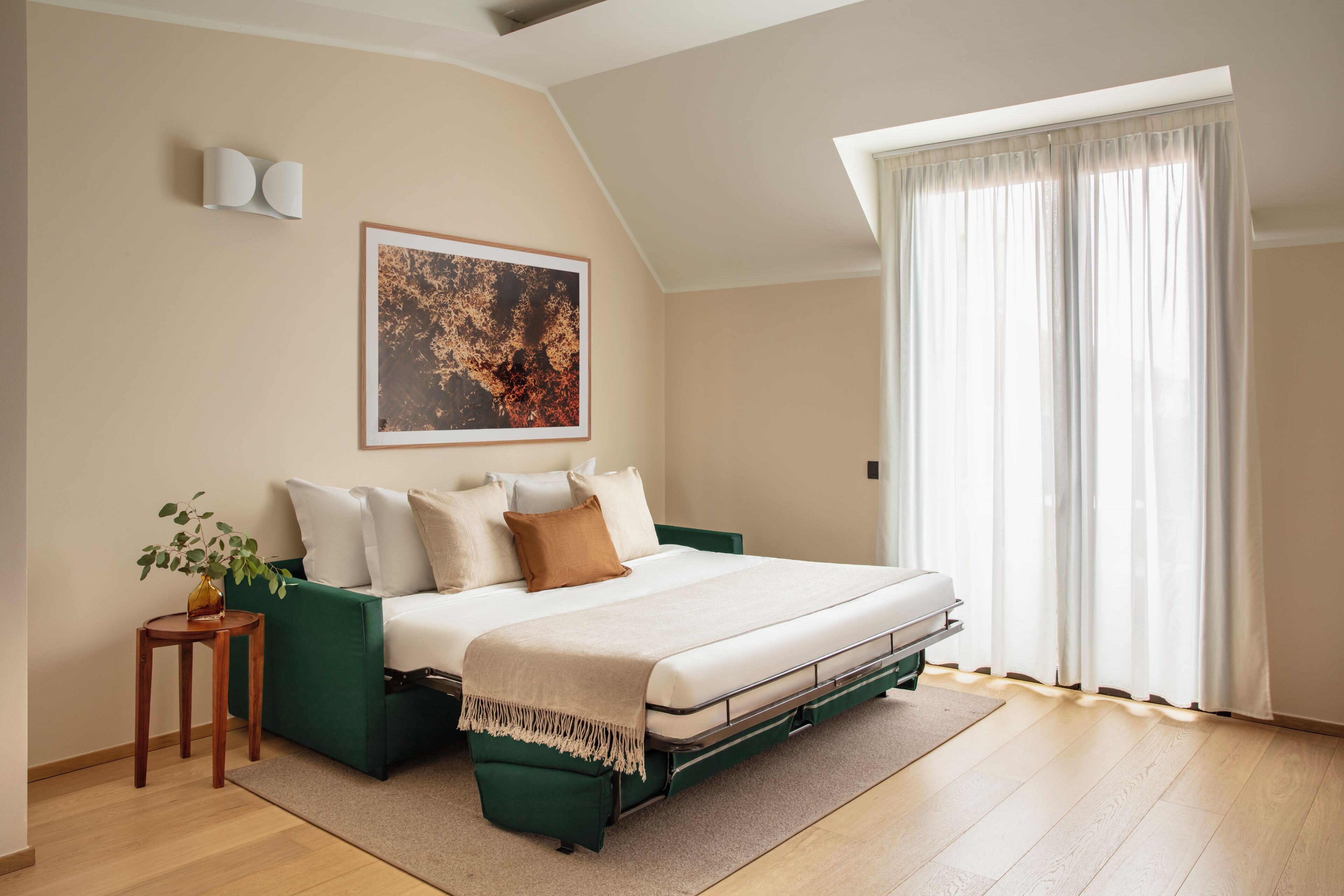 A bedroom with a bed, a green sofa, a small table with a plant, a painting on the wall, and a large window with curtains.