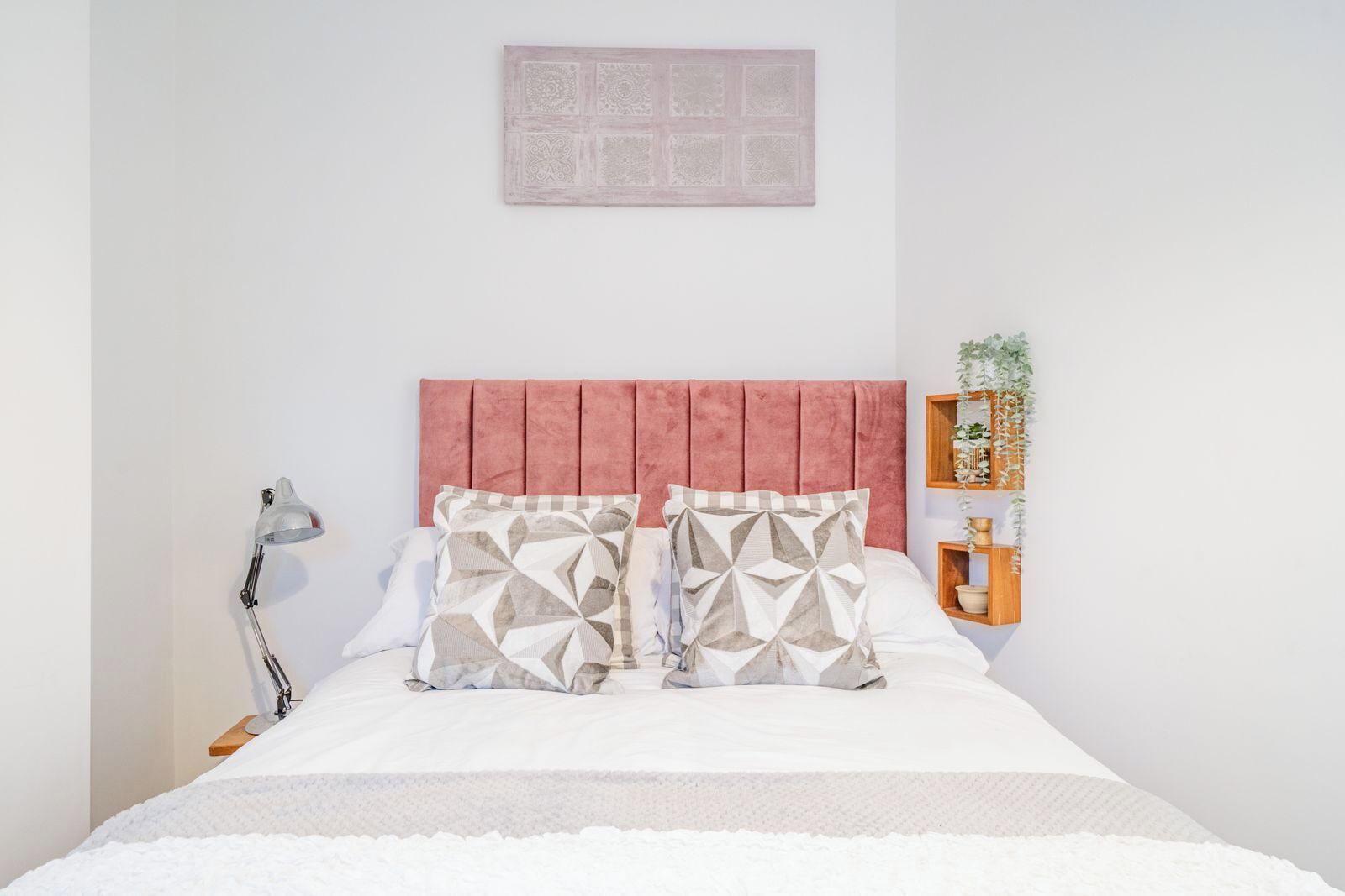 Ein Schlafzimmer mit Bett, Kopfstütze, Kissen, Nachttischlampe und einem an der Wand befestigten Regal mit einer Pflanze.