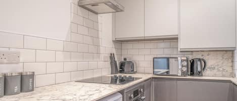 A modern kitchen with white tiles, a marble countertop, a built-in oven, a microwave, and a coffee machine.