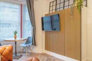 A modern room with a wooden slat wall, a flat-screen TV mounted above it, a round table with two chairs, and a potted plant on the window sill.