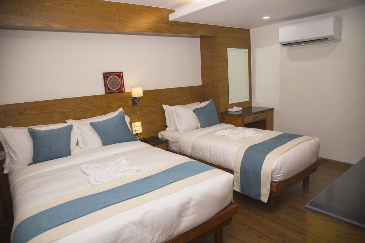Una habitación de hotel con dos camas, cabecera de madera, mesita de noche con lámpara, aire acondicionado y un espejo pequeño.