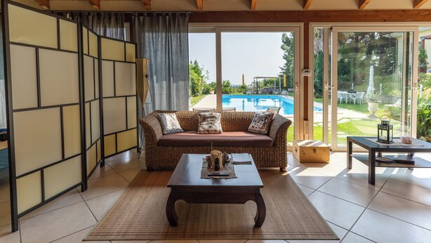 A room with a wooden ceiling, a sofa with cushions, a table with a mat, and a view of a swimming pool through glass doors.