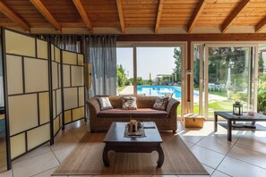 Una habitación con techo de madera, un sofá con cojines, una mesa con un felpudo y vistas a una piscina a través de puertas de vidrio.
