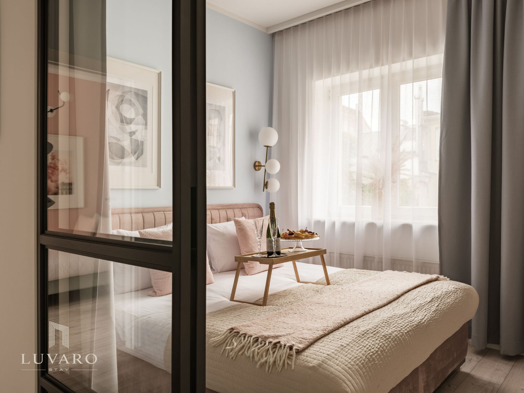 A bedroom with a bed, a bedside table with a bottle and glasses, a lamp, and a window with curtains.